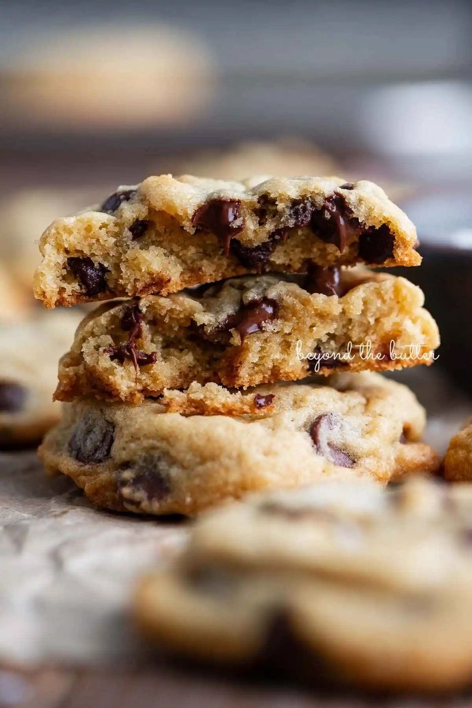 Chocolate chip cookies randomly placed on crumpled parchment paper with two cookies stacked on top of the other. The top cookie is split in half to see the soft-centered center.