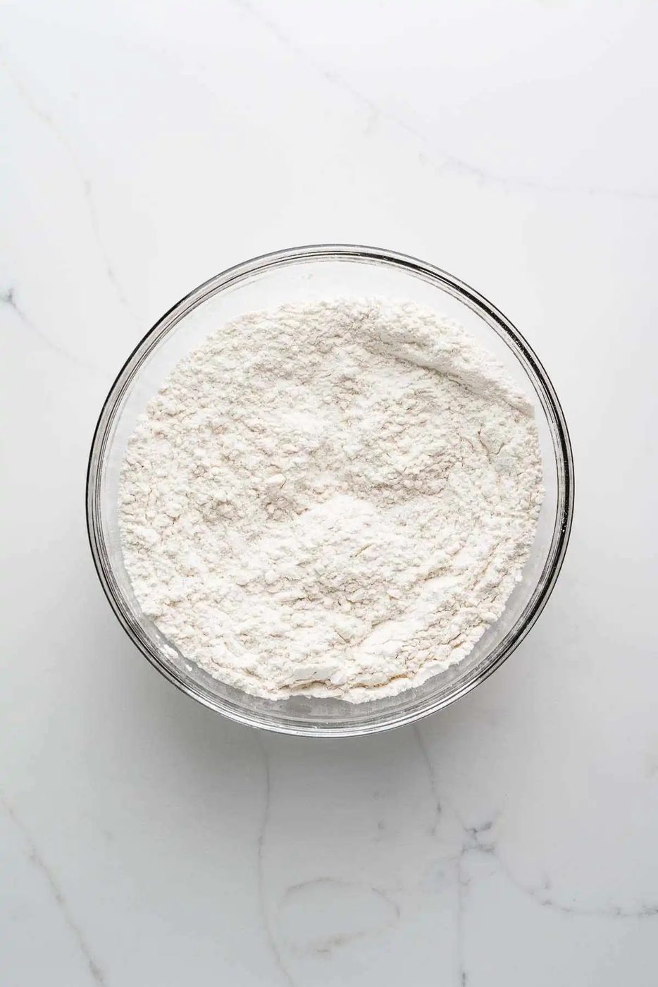 Whisked all purpose flour, baking soda, and vanilla flavored instant pudding mix in a large glass mixing bowl on a white marbled background.