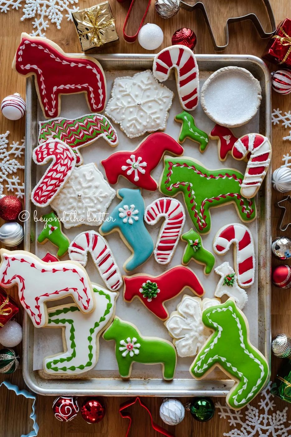 Randomly placed decorated cut out sugar cookies on a parchment lined baking sheet with ornaments and cookie cutters around it | All Images © Sovuh