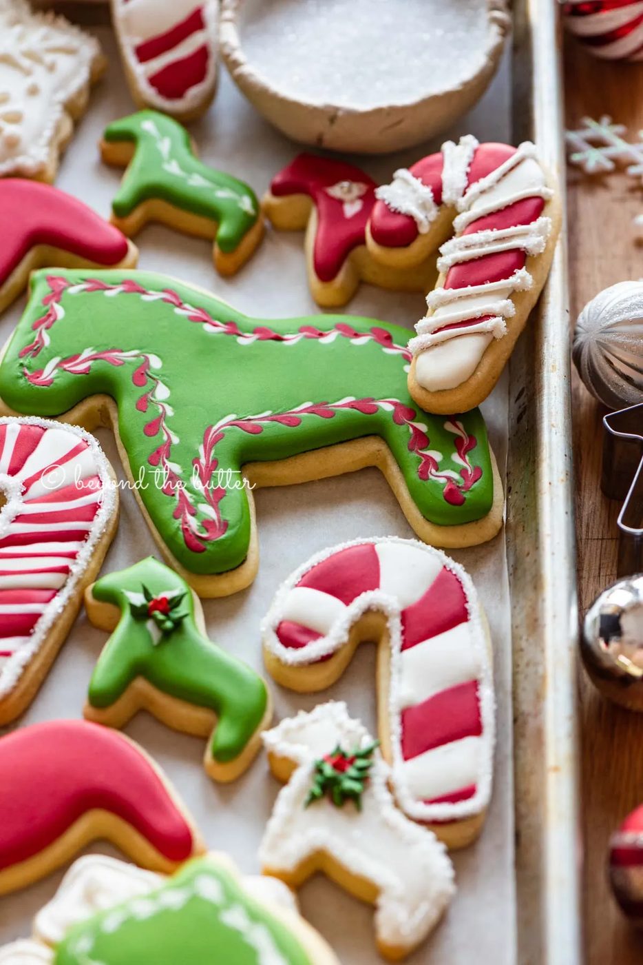 Randomly placed decorated cut out sugar cookies on a parchment lined baking sheet with ornaments and cookie cutters around it | All Images © Sovuh