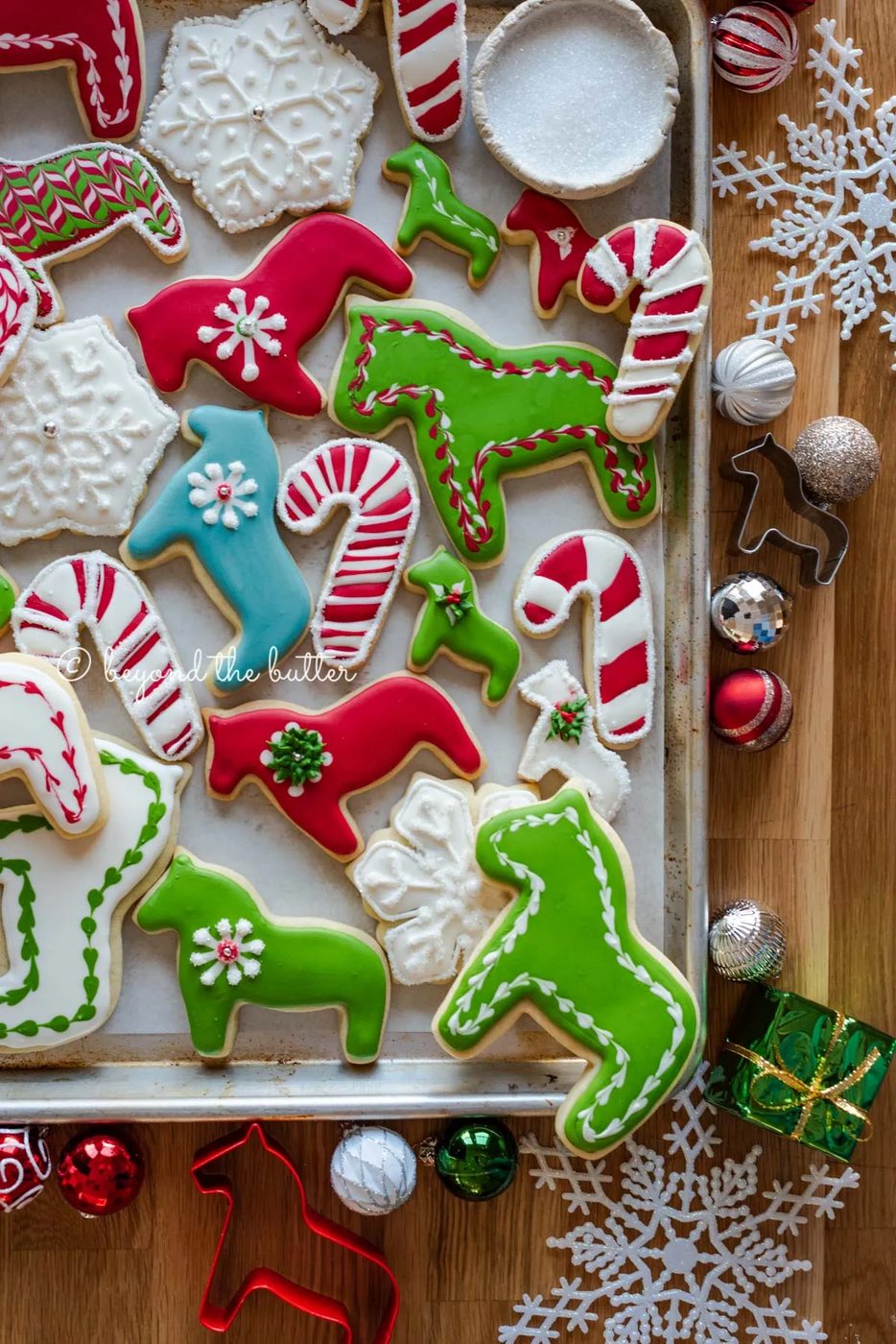 Randomly placed decorated cut out sugar cookies on a parchment lined baking sheet with ornaments and cookie cutters around it | All Images © Sovuh