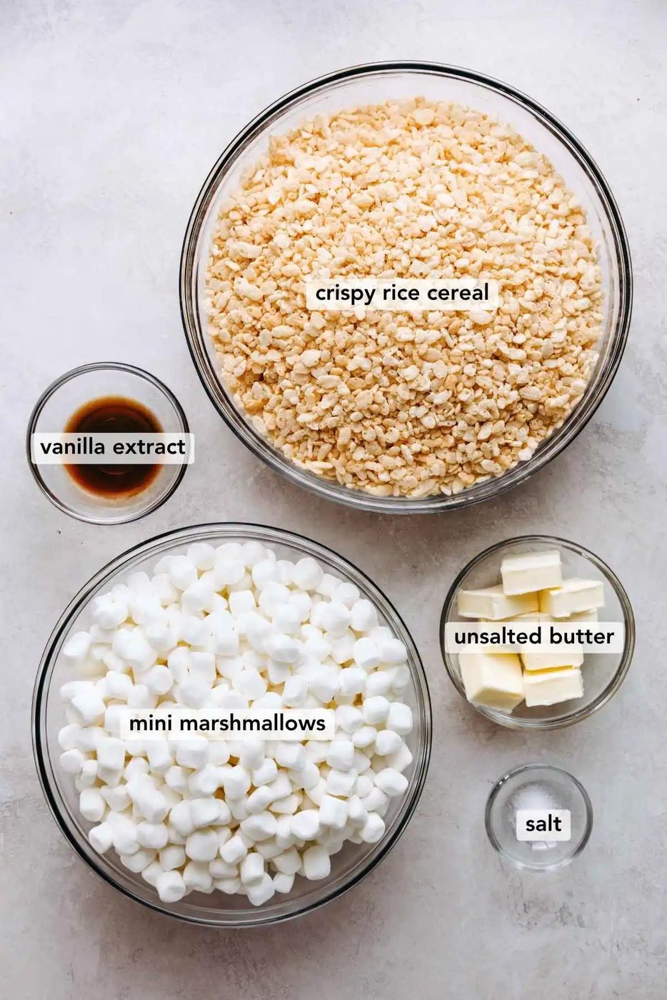 Ingredients in various size plates and bowls labeled as rice krispies cereal, vanilla extract, mini marshmallows, unsalted butter, and salt on a white marbled background.