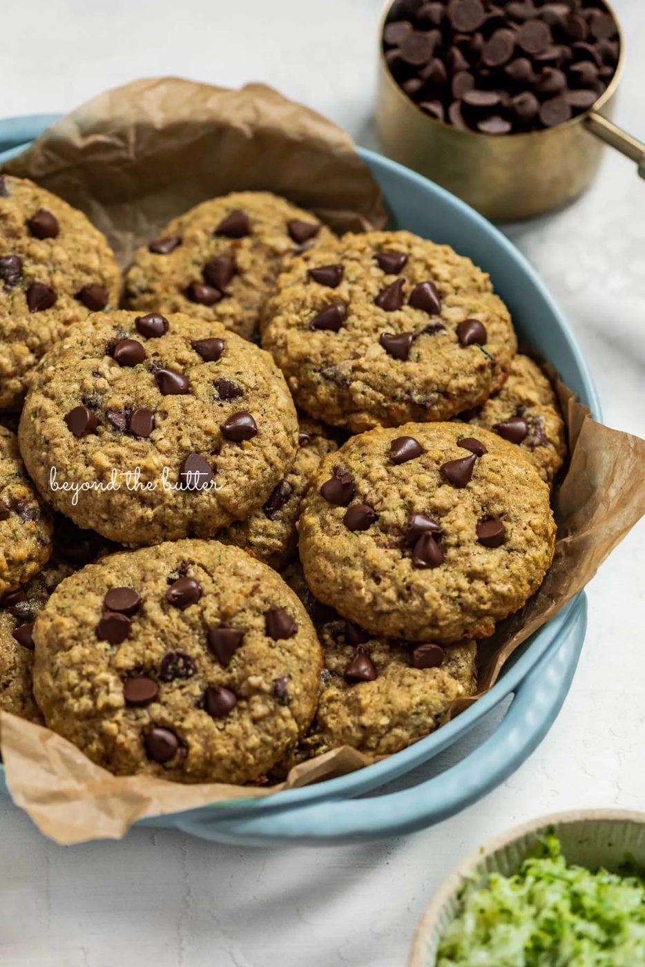 Baked zucchini chocolate chip oatmeal cookies in a parchment paper lined blue serving dish with a measuring cup of chocolate chips and a bowl of shredded zucchini nearby | © Sovuh