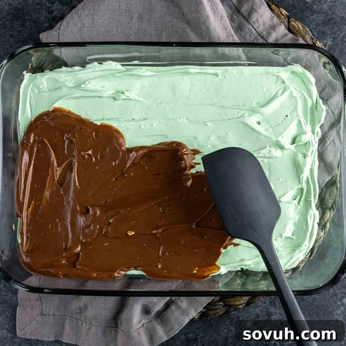 A spatula smoothing out the chocolate pudding layer over the vibrant green cream cheese layer in a glass baking dish.