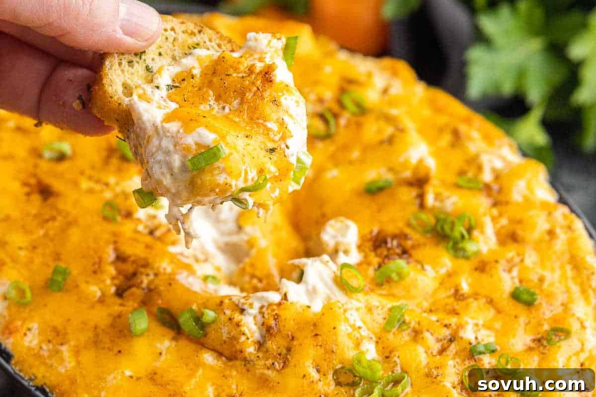 Close-up of a hand dipping a piece of bread into a creamy, baked cheese dip topped with chopped green onions.