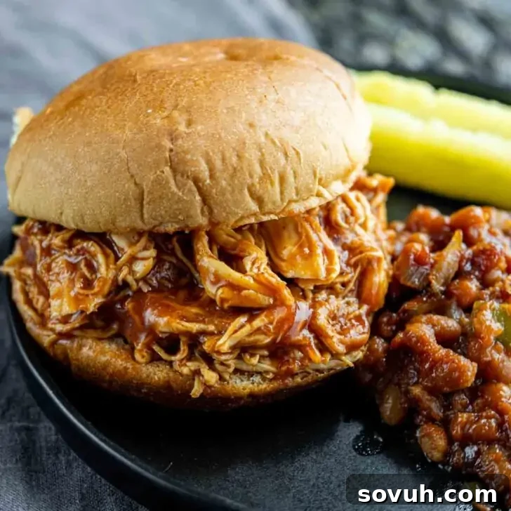 A pulled chicken sandwich with barbecue sauce is on a black plate, accompanied by baked beans and a few pickle spears.