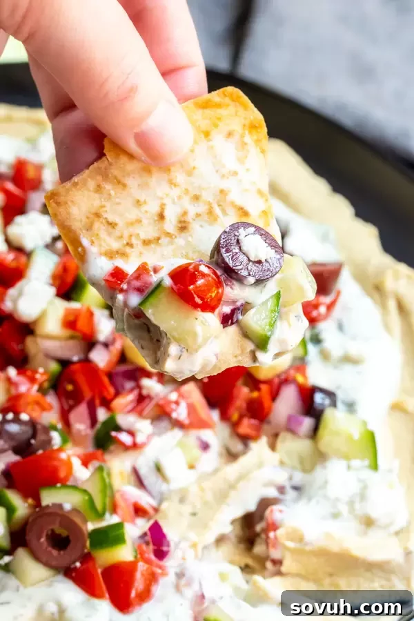 Hand holding a pita chip with a generous scoop of 5 Layer Greek Dip