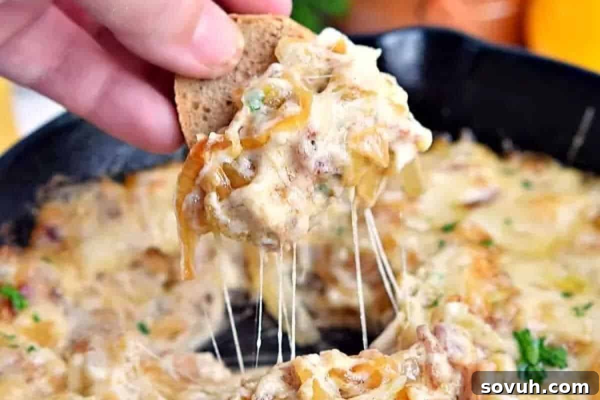 A close-up shot of a golden cracker being dipped into a rich, cheesy caramelized onion and bacon skillet dip, with tantalizing melted cheese stretching from the skillet.