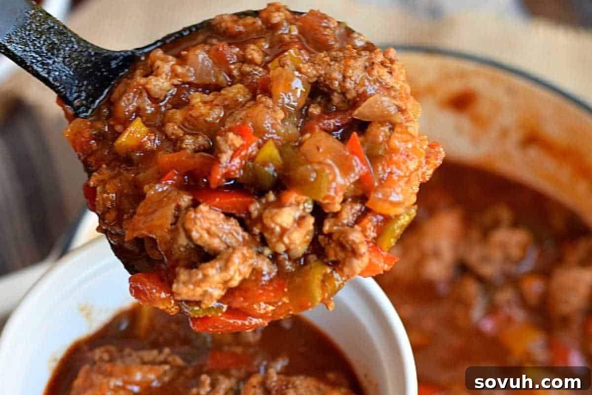 A ladle brimming with hearty Pale Ale Chili, rich with ground beef, ground pork, tomatoes, and peppers, poised over a full bowl of the comforting stew.