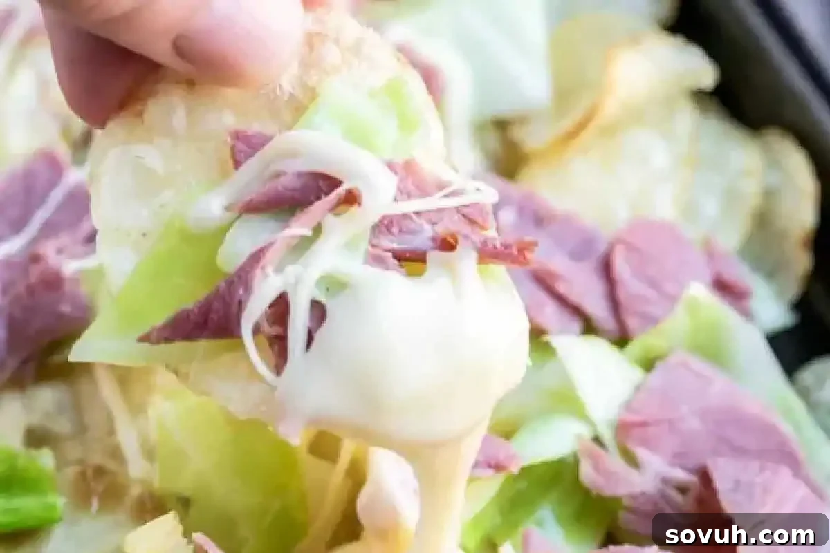 A hand holding a golden, crispy potato chip generously topped with melted beer cheese sauce, tender corned beef, and fresh cabbage, showcasing the unique twist of Irish Nachos.