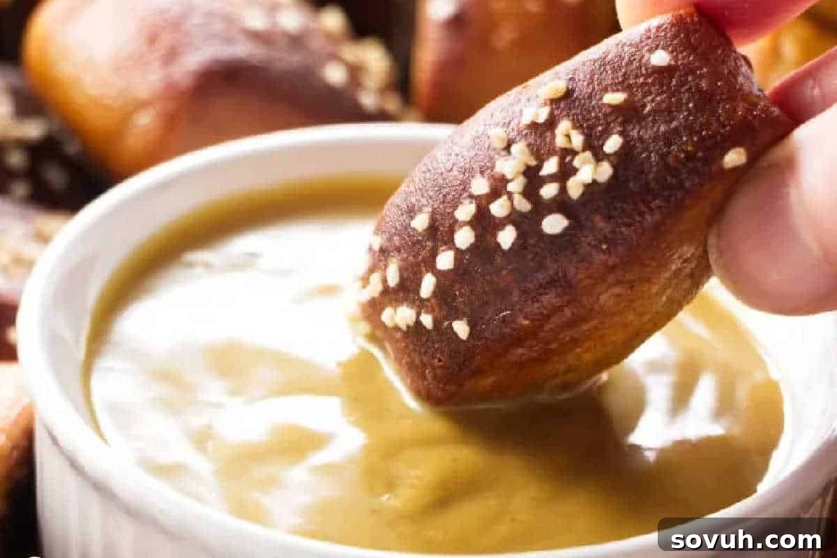 A freshly baked, sesame seed-coated beer pretzel bite being dipped into a small bowl of tangy mustard sauce, highlighting its golden exterior.