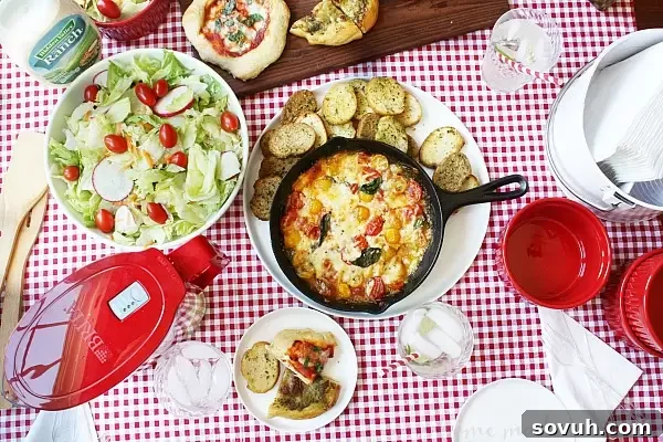 A beautifully arranged tablescape for a casual adult lunch party, featuring individual pizzas, salad, and the centerpiece, a warm Baked Caprese Dip.