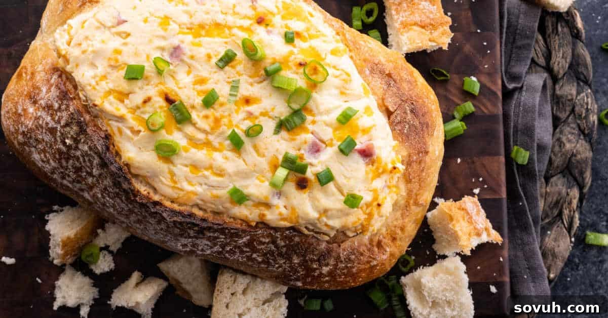 Cheese dip in a bread bowl topped with chopped green onions, surrounded by bread pieces on a wooden board.