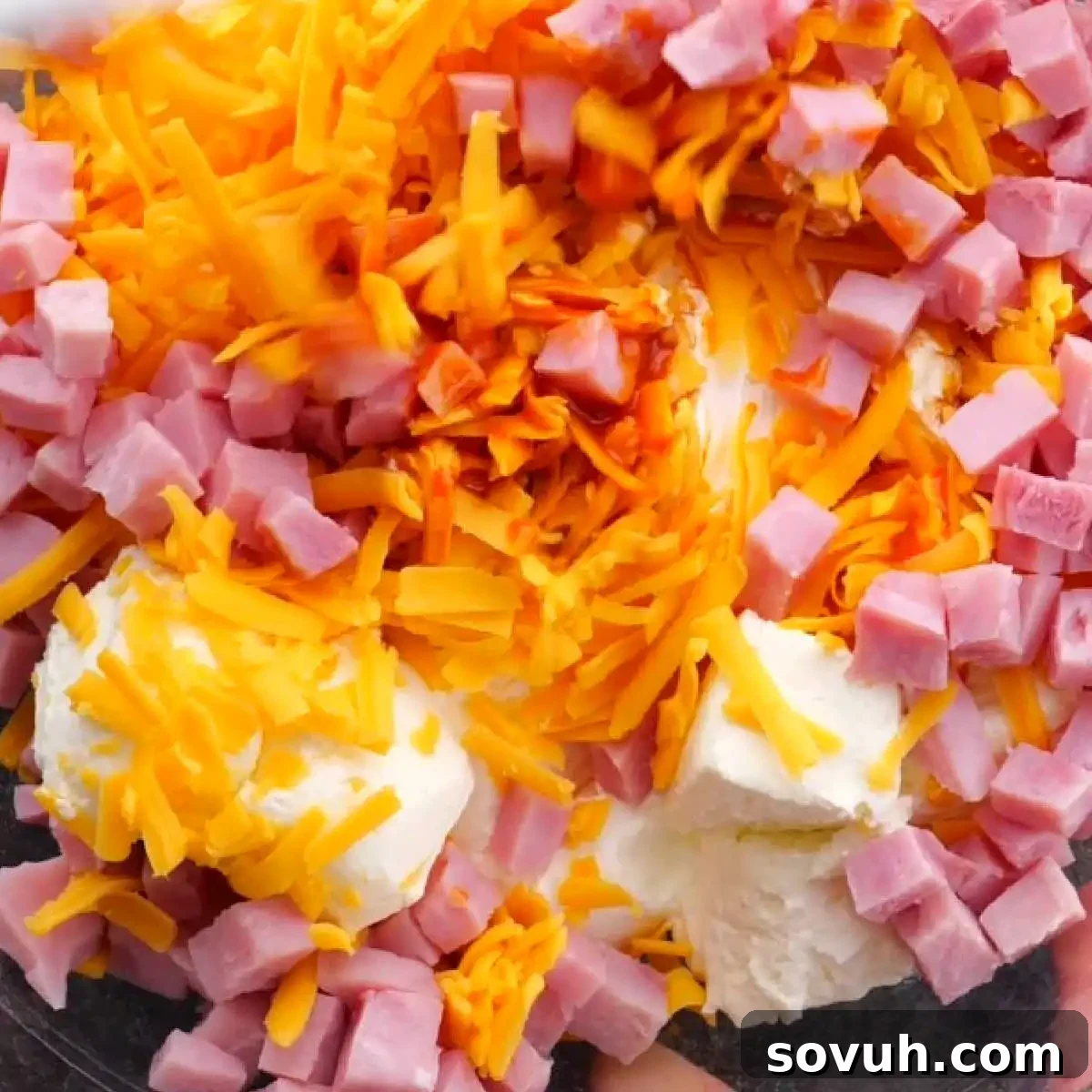 A bowl filled with chopped ham, shredded cheddar cheese, and dollops of cream cheese.