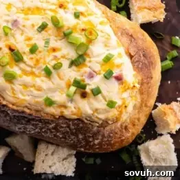 A loaf of bread hollowed and filled with creamy cheese dip, topped with chopped green onions, surrounded by bread pieces on a wooden surface.