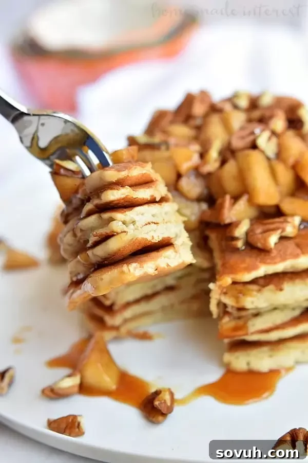 A close-up of a fork piercing a stack of Caramel Apple Pecan Pancakes, revealing their fluffy texture and the delicious layers of baked apples, pecans, and caramel sauce.