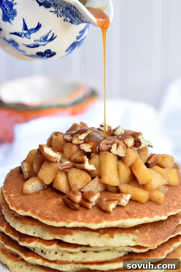 Close-up shot of rich caramel maple syrup being poured over a stack of Caramel Apple Pecan Pancakes, highlighting the golden brown pancakes, baked apples, and chopped pecans.