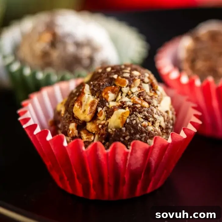 A chocolate truffle coated with chopped nuts sits in a red paper cup, with other truffles blurred in the background.