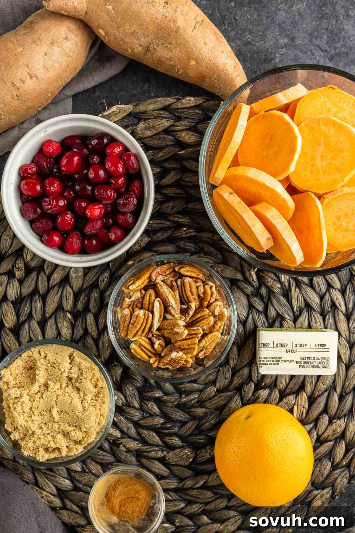 Various ingredients for Cranberry Sweet Potato Casserole laid out on a wooden board: sliced sweet potatoes, fresh cranberries, an orange, butter, cinnamon, brown sugar, and pecans.