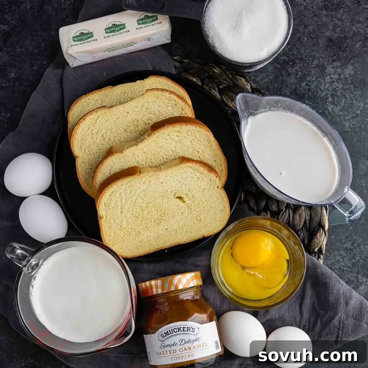 Ingredients for a recipe are arranged: bread slices, butter, sugar, milk, cream, eggs in a bowl, and a jar of salted caramel topping on a dark surface.