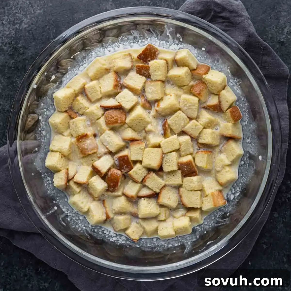 A glass bowl filled with bread cubes soaking in a liquid mixture, placed on a dark surface with a gray cloth underneath.