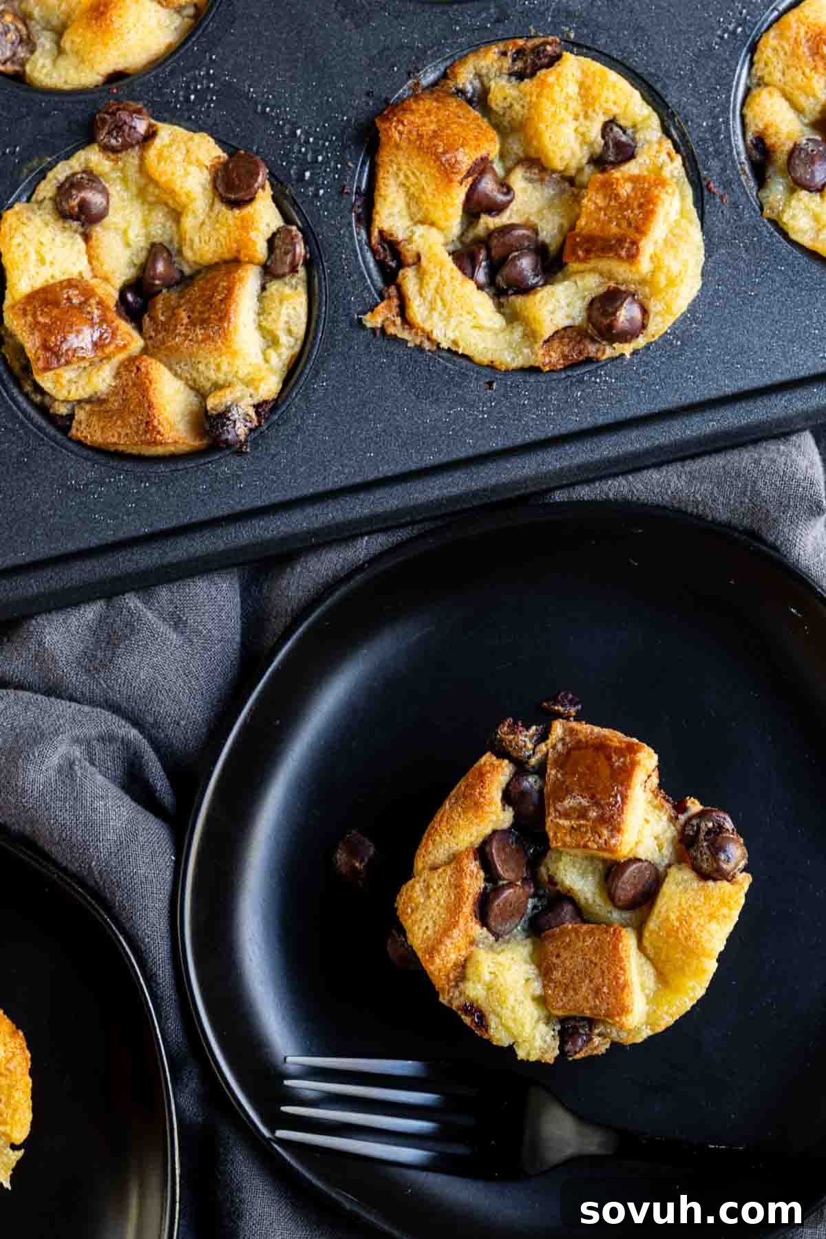 Chocolate chip bread pudding muffins in a black muffin tin and plates, with a dark cloth underneath.