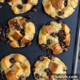 Six freshly baked muffins in a dark baking tray are topped with chocolate chips and chunks of bread.