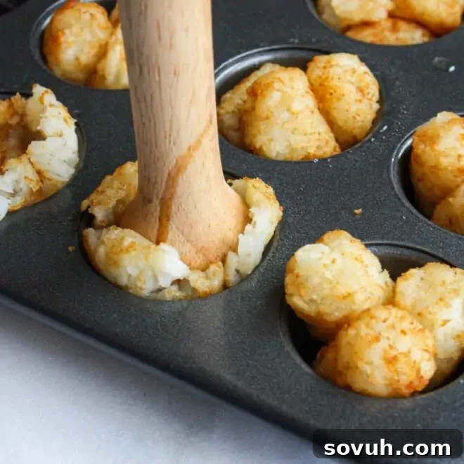 A muffin tin filled with partially baked tater tots, ready to be pressed into cups for cheeseburger filling.