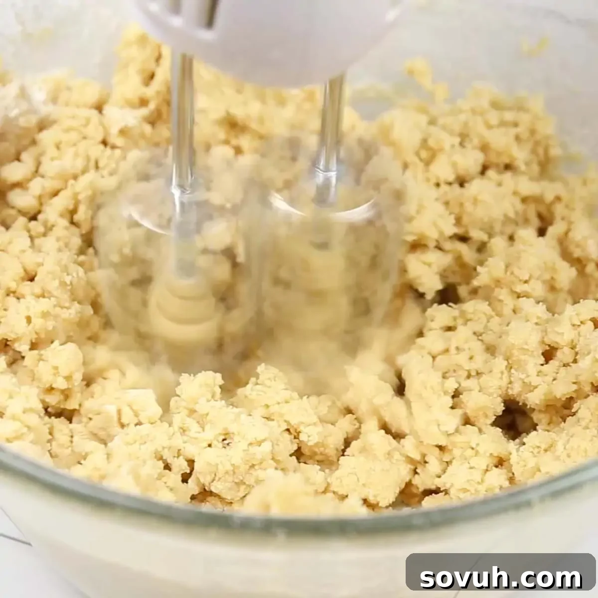 A hand mixer skillfully combining sugar, brown sugar, and softened butter in a large mixing bowl, creating a creamy base for the chocolate chip cookie dough layer of the brownie cake.