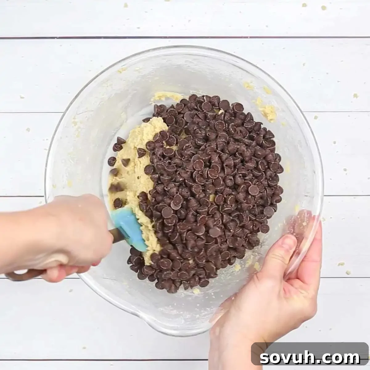 A hand gently scooping a generous handful of semi-sweet chocolate chips into the creamy, spreadable chocolate chip cookie dough, ready to be folded in for the brownie cake layers.