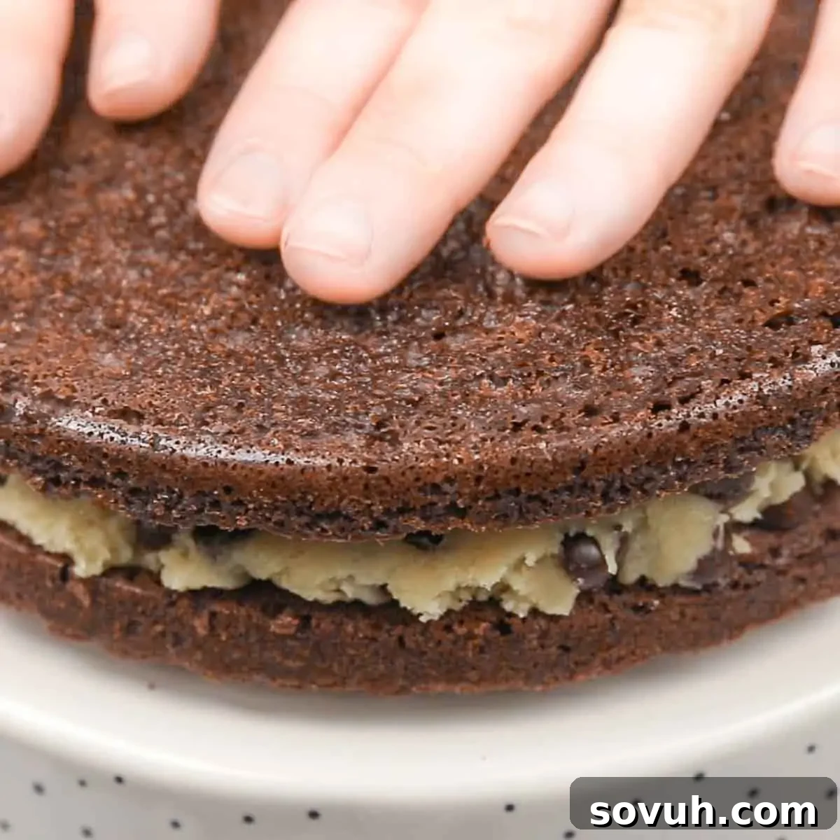 A hand gently patting down the top layer of a multi-tiered brownie cake, ensuring an even surface before the final chocolate ganache and brownie chunk toppings are applied.