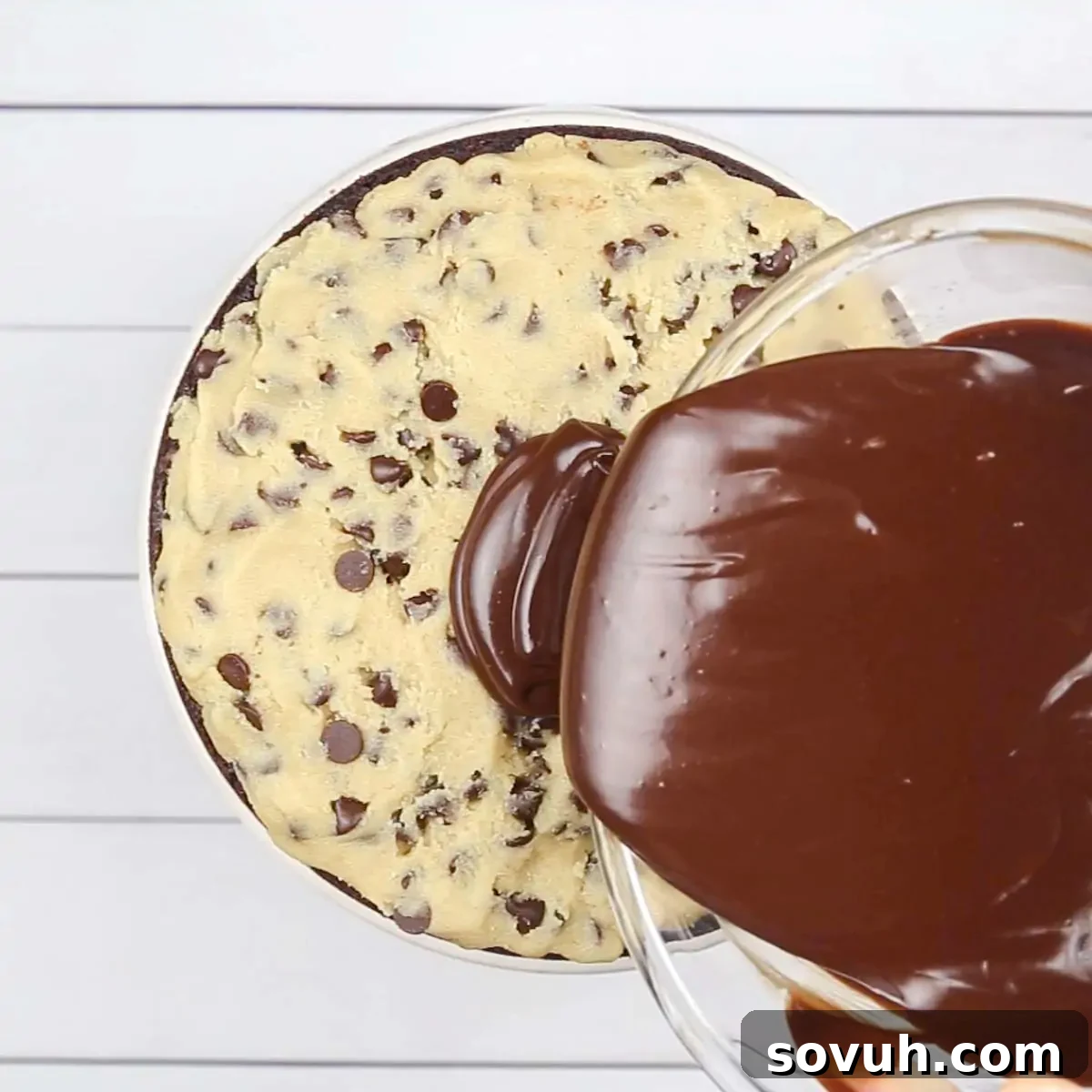 A stream of glossy, dark chocolate ganache being drizzled artfully over the top layer of chocolate chip cookie dough on a layered brownie cake, creating a decadent finish.