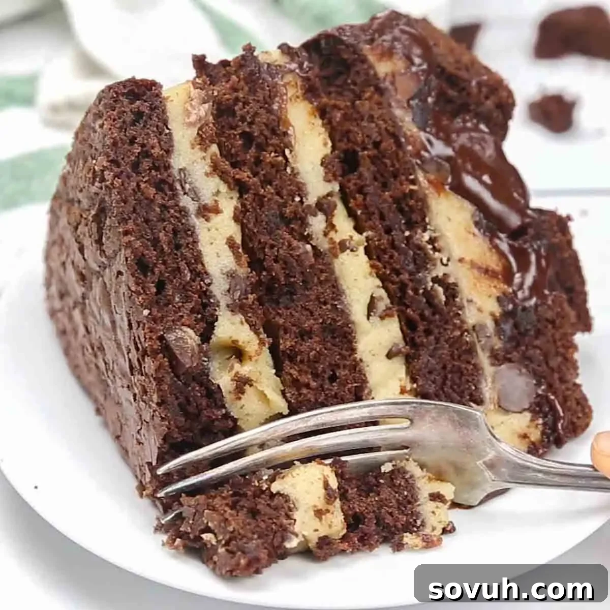 A perfect slice of layered brownie cake, showcasing the fudgy brownie, creamy cookie dough, and luscious ganache, served on a white plate with a silver fork.