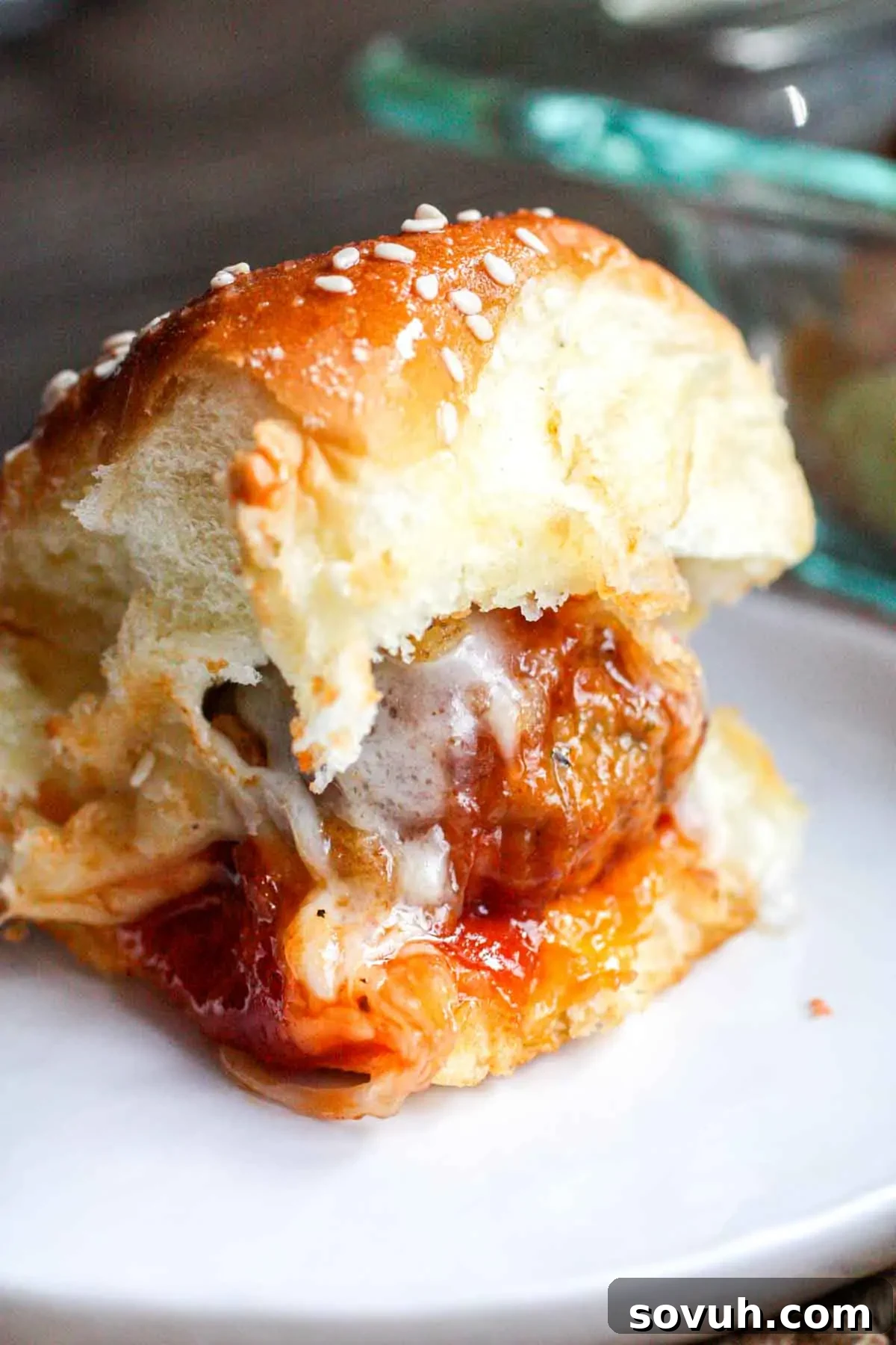 Effortless Tangy Meatball Sliders 3 A bitten meatball slider with marinara sauce and melted cheese on a sesame-seed bun sits on a white plate.