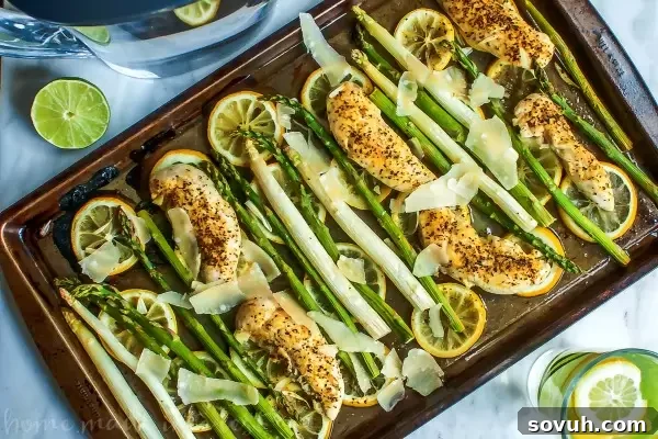 Raw chicken tenders and fresh asparagus laid out on a sheet pan with lemon slices, ready for seasoning