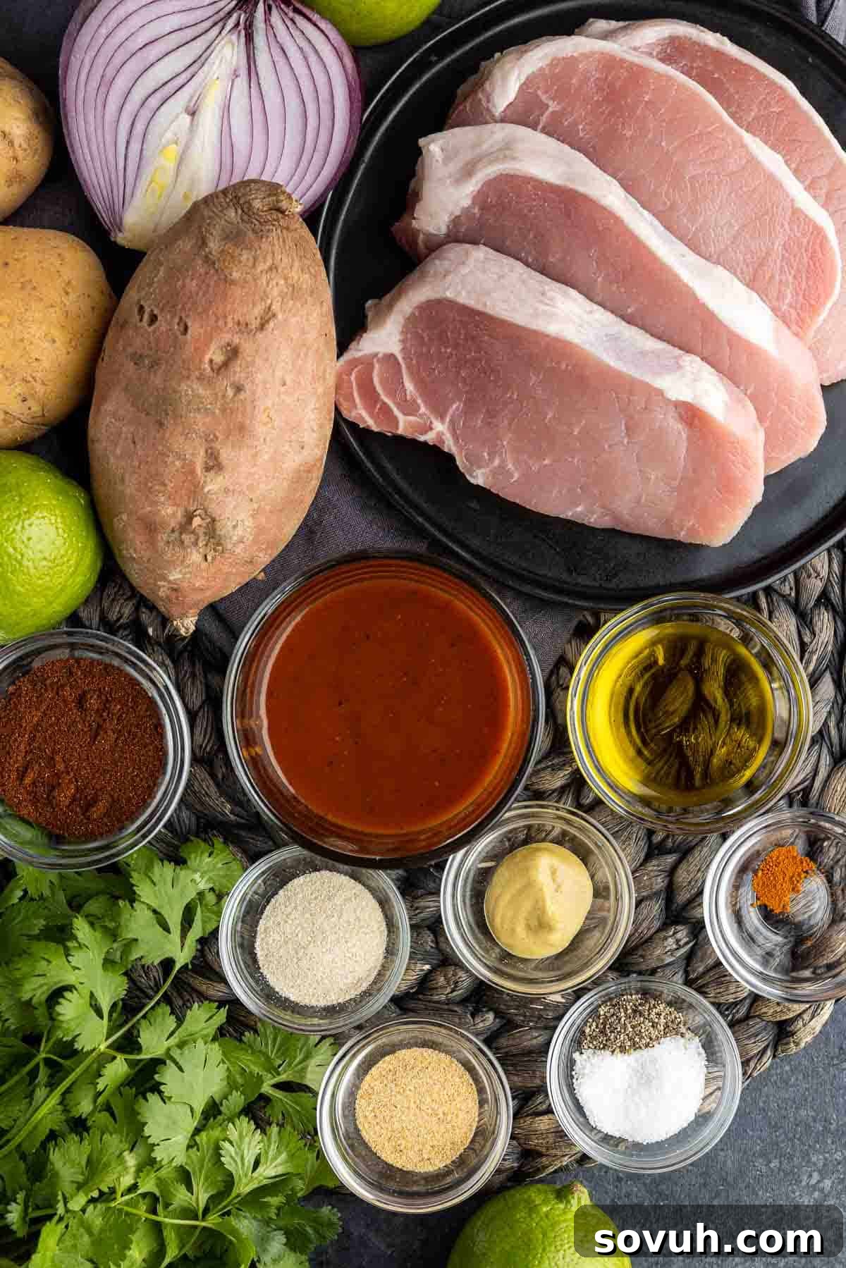 Assorted ingredients laid out for Sheet Pan Spicy BBQ Pork Chops and Potatoes, including pork chops, various spices, olive oil, different types of potatoes, red onion, BBQ sauce, Dijon mustard, lime, and fresh cilantro.