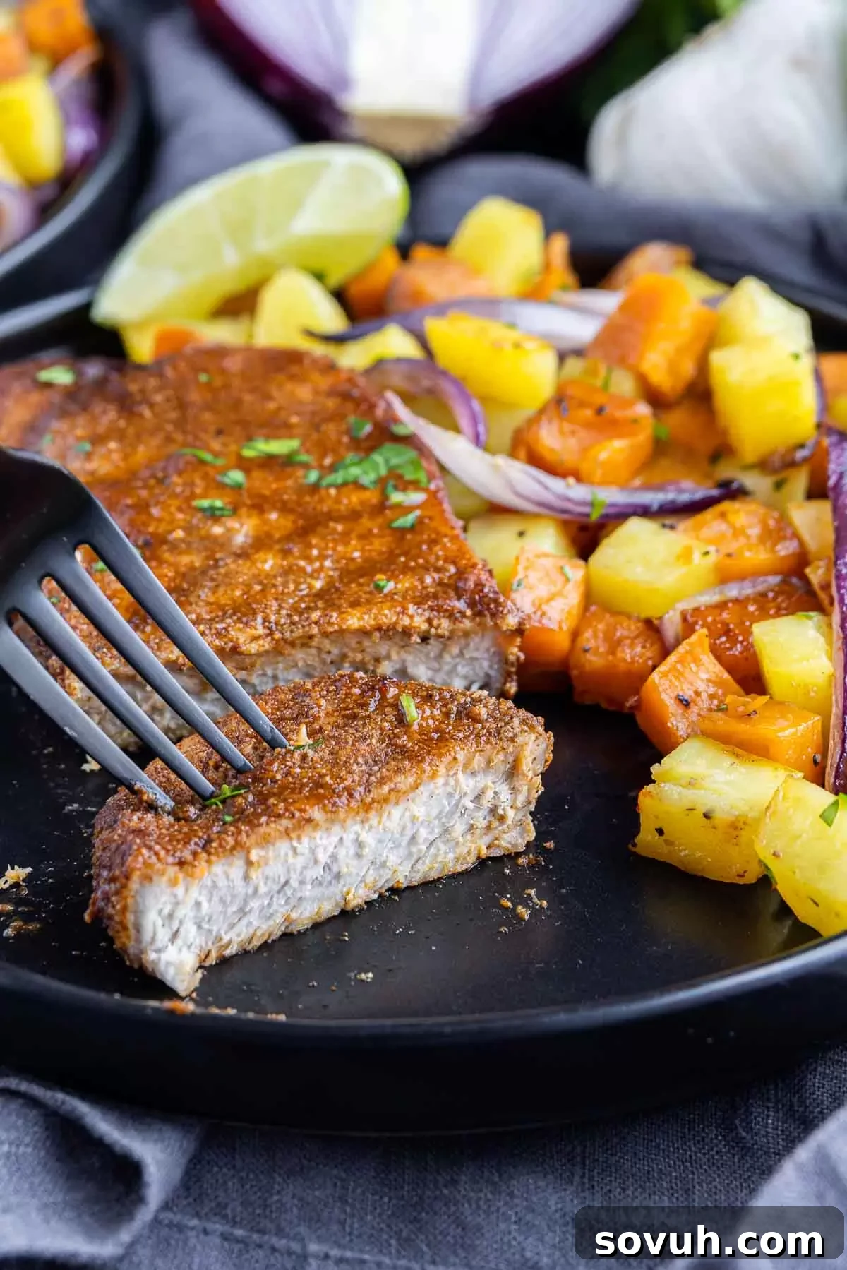 A hand cutting into a perfectly cooked Sheet Pan Spicy BBQ Pork Chop, revealing its tender, juicy interior, alongside roasted potatoes on a sheet pan.
