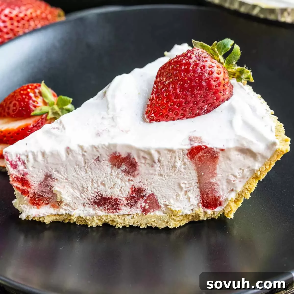 Slice of Frozen Strawberry Pie on a black plate with fresh strawberries on top, next to the full pie
