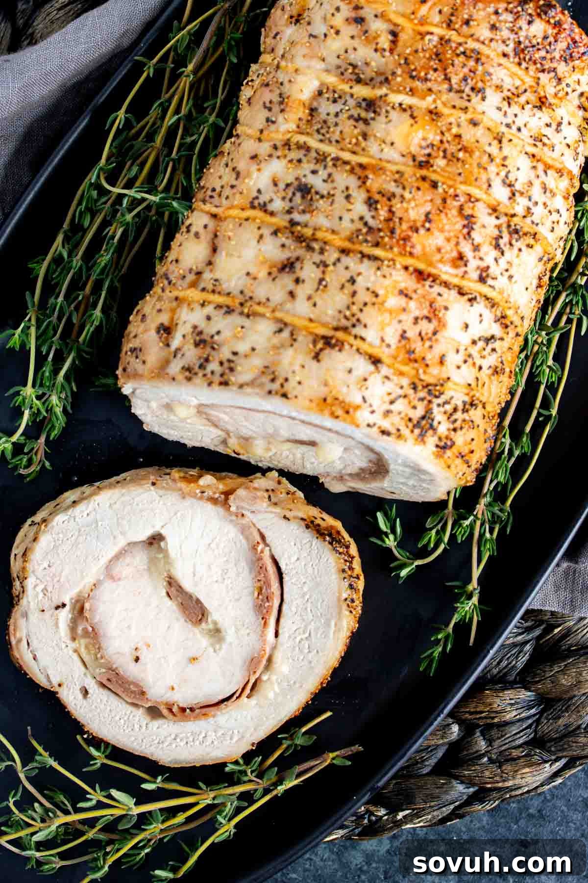 Another view of the Provolone and Prosciutto Stuffed Pork Loin on a black plate, artfully arranged with fresh thyme sprigs. This shows the beautiful golden crust and tender interior of this delicious baked pork loin.