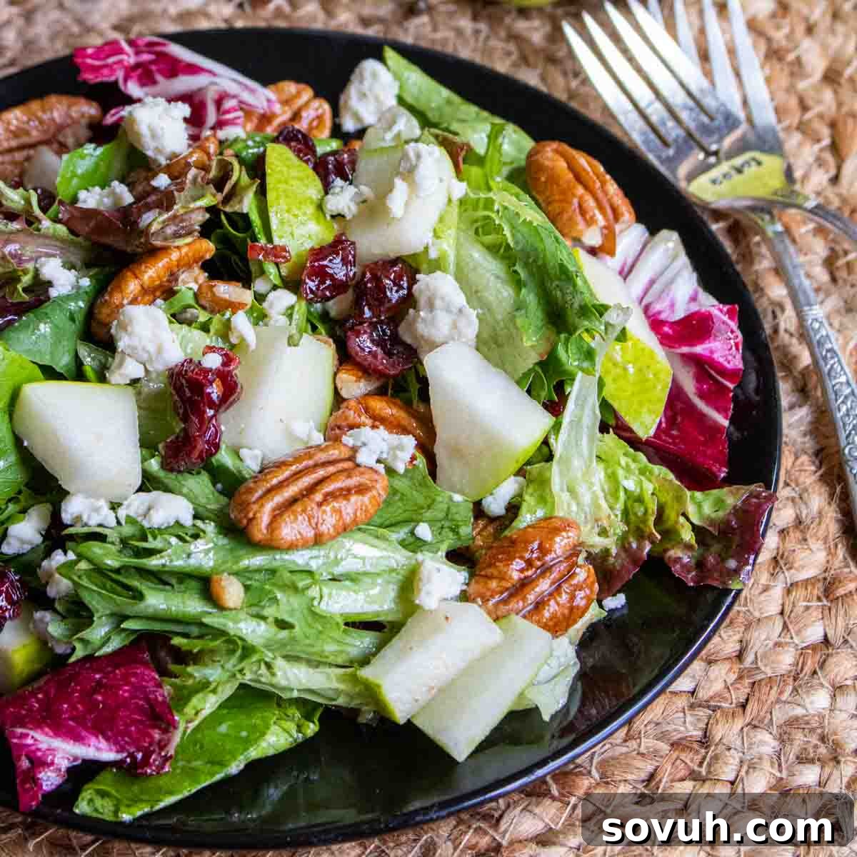 A vibrant winter salad with pears, cranberries, and pecans on a black plate, with forks ready to serve.