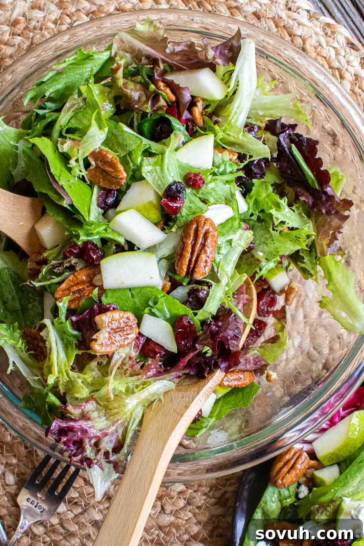 A large glass bowl filled with tossed winter salad, featuring mixed greens, pears, cranberries, and pecans, with wooden serving spoons.