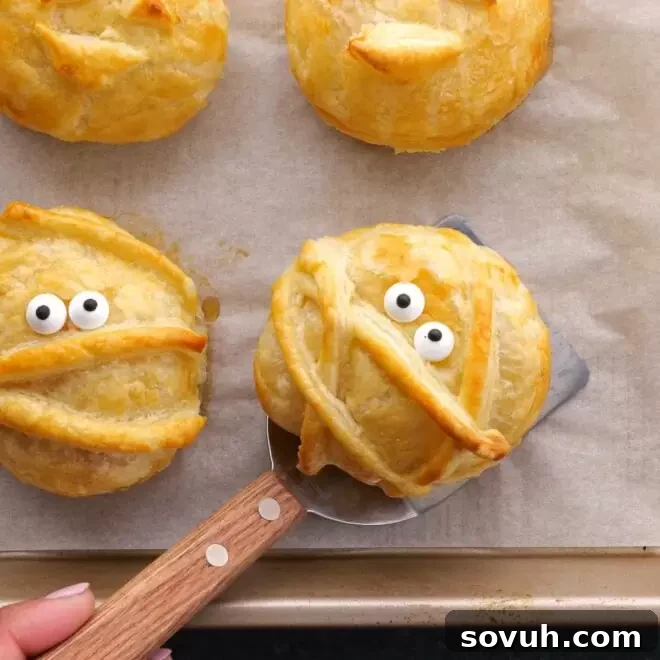 A hand holding a warm Halloween Baked Cheese mummy, showcasing its golden puff pastry and edible eyes.