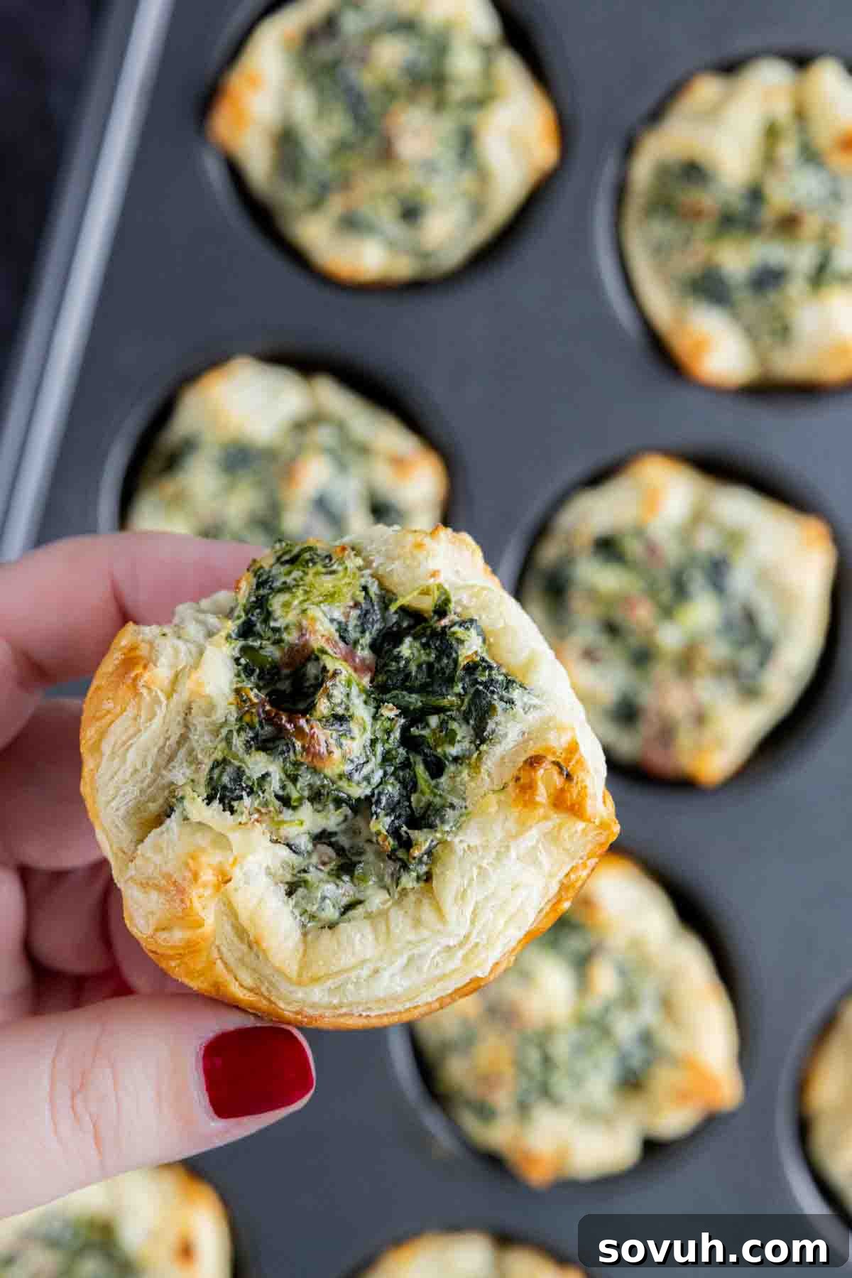 A hand gently lifting a perfectly golden and crisp spinach puff pastry from a muffin tin, highlighting its flaky texture.