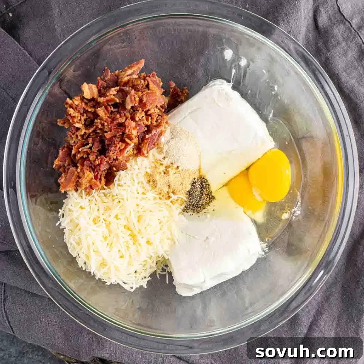 A large mixing bowl containing softened cream cheese, two eggs, shredded Parmesan, and finely chopped cooked bacon, ready to be combined for the spinach puff filling.