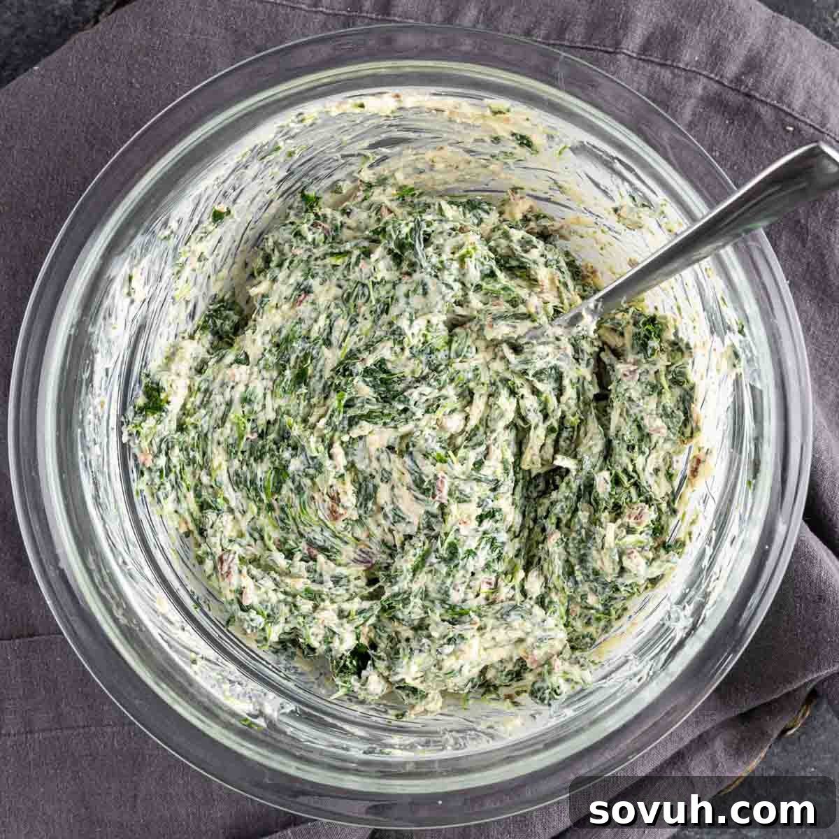 A bowl filled with the creamy, cheesy spinach and bacon mixture, with a spoon resting inside, ready for the puff pastry.