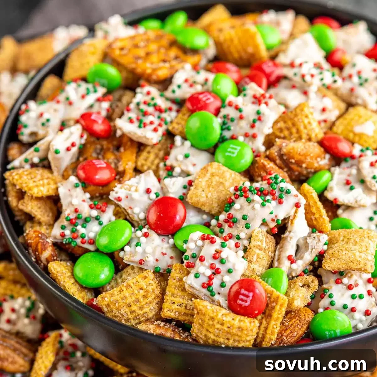 Close up of a black bowl filled with sweet and savory Christmas Chex Mix, featuring cereals, pretzels, nuts, and red and green candies.