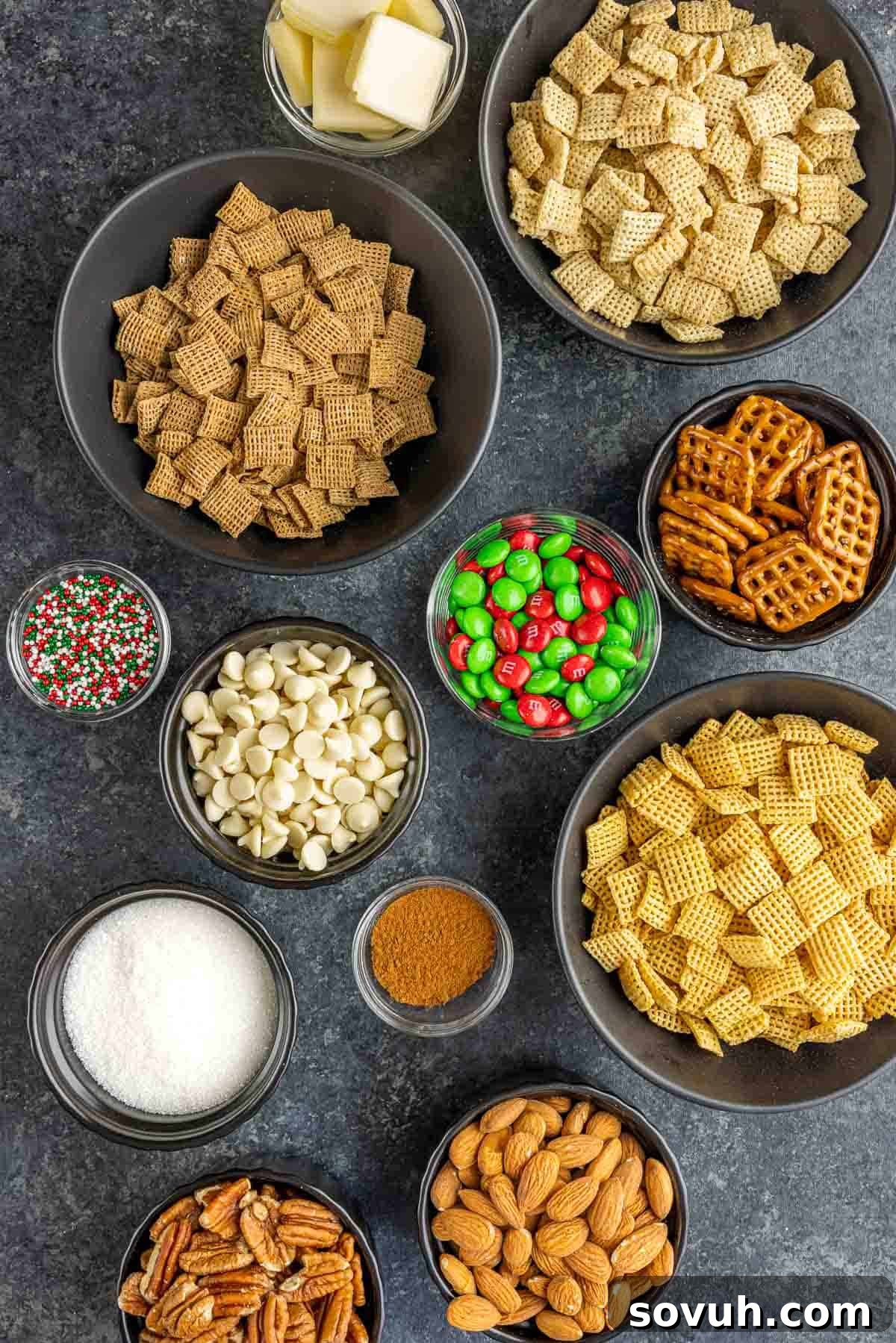 Assortment of ingredients for Christmas Chex Mix laid out on a white surface, including three types of Chex cereal, nuts, pretzels, sprinkles, M&Ms, white chocolate chips, butter, sugar, and cinnamon.