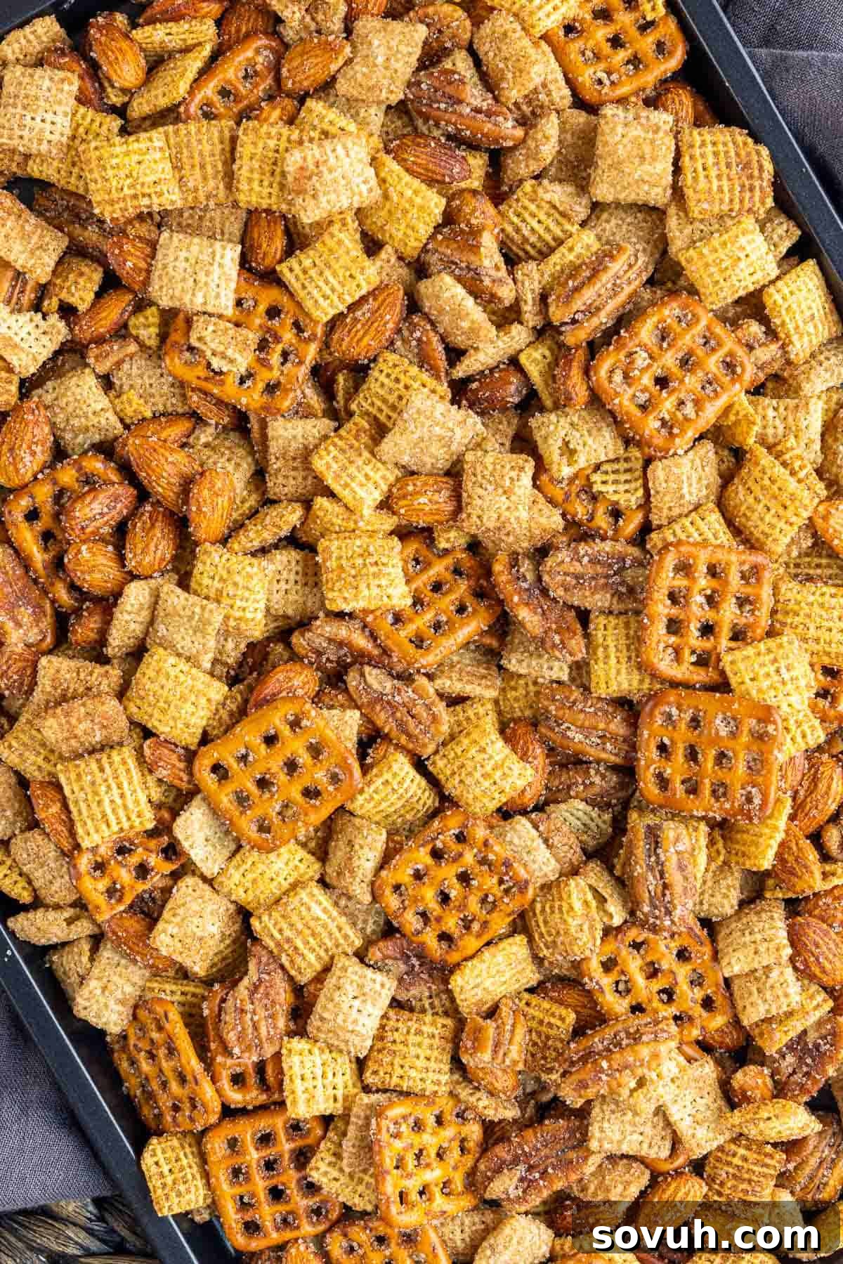 A baking sheet with corn and wheat Chex cereal, almonds, pecans, and mini pretzels, coated with butter and cinnamon sugar, ready for microwaving.
