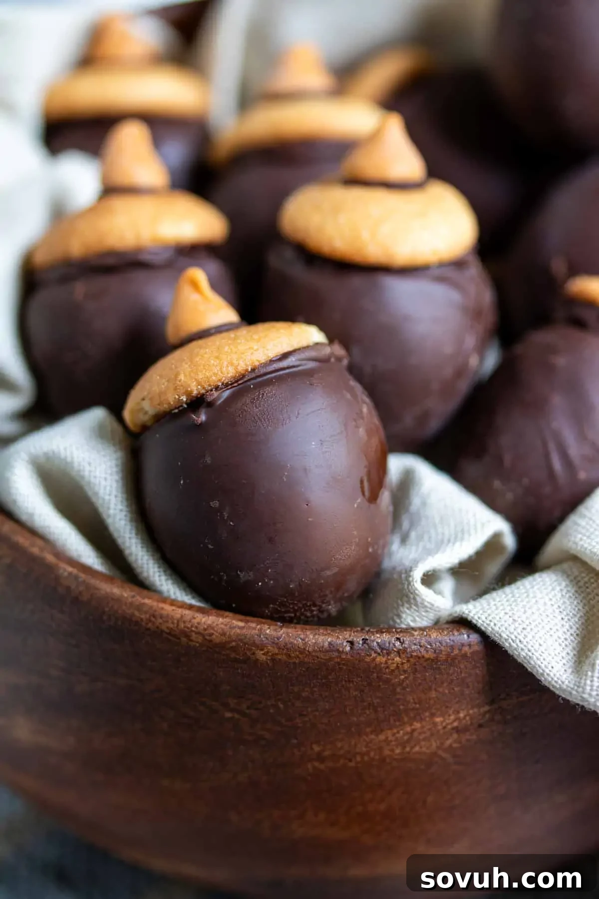 A wooden bowl filled with chocolate-covered acorn-shaped treats with a cookie and chocolate chip on top, resting on a cloth napkin.