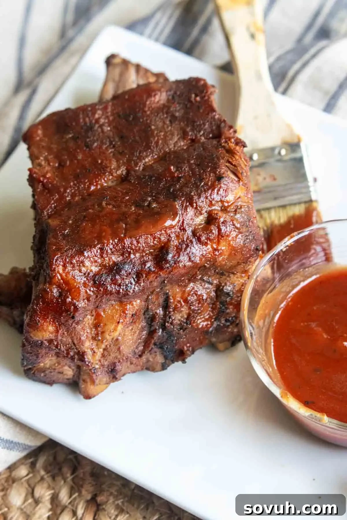 A serving of Slow Cooker Ribs sits on a white plate next to a small bowl of barbecue sauce and a basting brush.